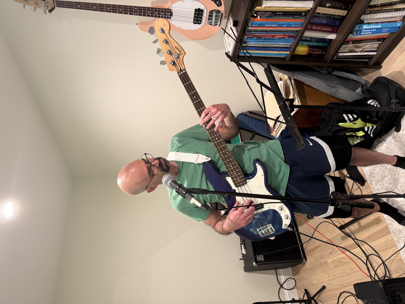 Adult student playing electric bass during an in-person lesson, music stand and bookshelves of sheet music in background