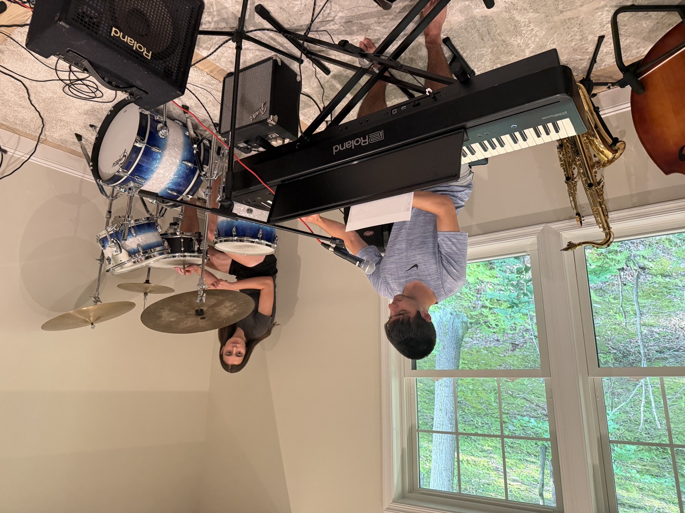 Two students rehearsing together — one on drums, one at a Roland keyboard — with saxophone and upright bass visible in the background